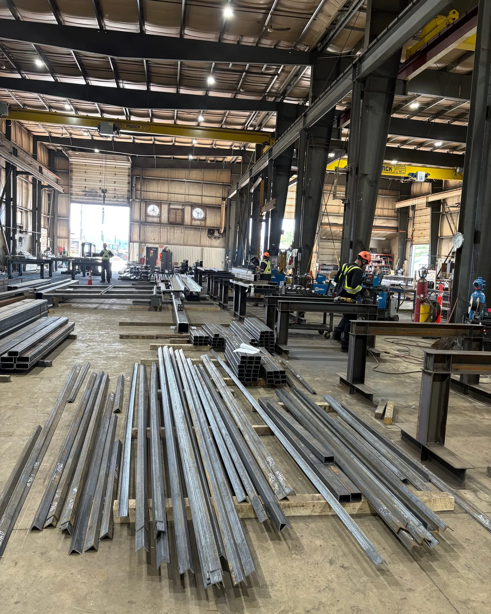 Steel angles and tubes laid out on the shop floor in front of welding stations, with workers in hard hats at the benches and an overhead crane above.