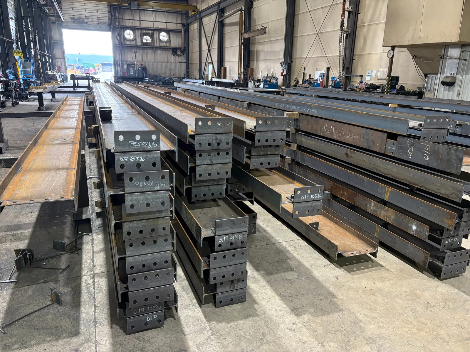 Rows of fabricated wide-flange steel beams stacked in the shop, their drilled end connection plates marked with piece numbers.
