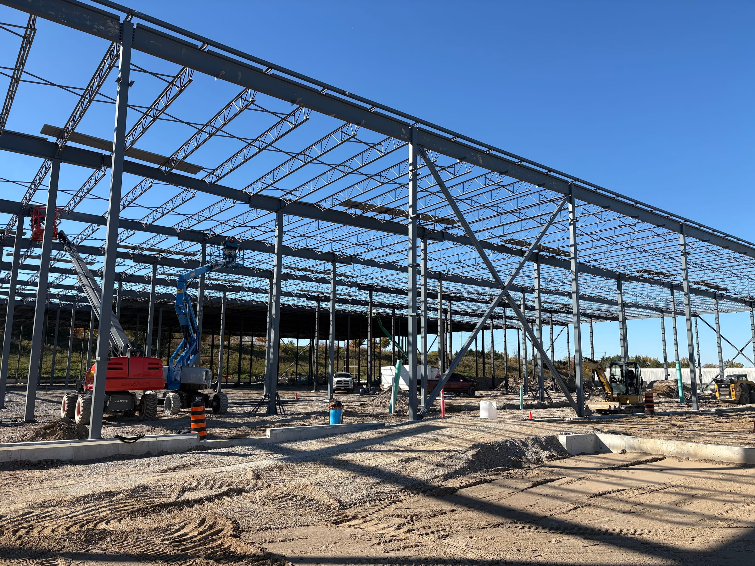 Looking up through a grid of steel columns and open-web roof joists against a deep blue sky, with boom lifts working among the columns.