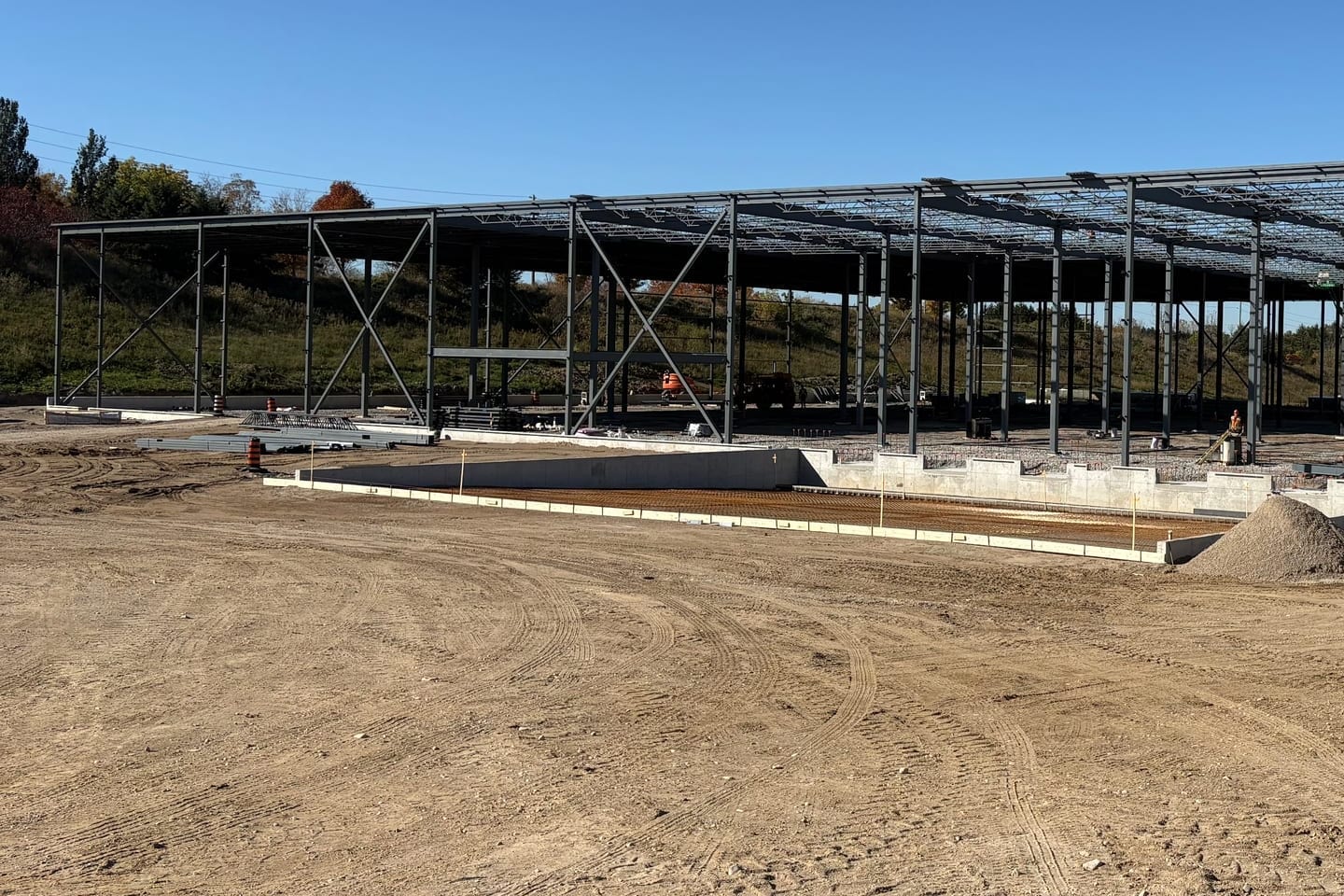 The long side of the braced steel frame across a graded site, with roof joists along the top and a concrete foundation wall in the foreground.