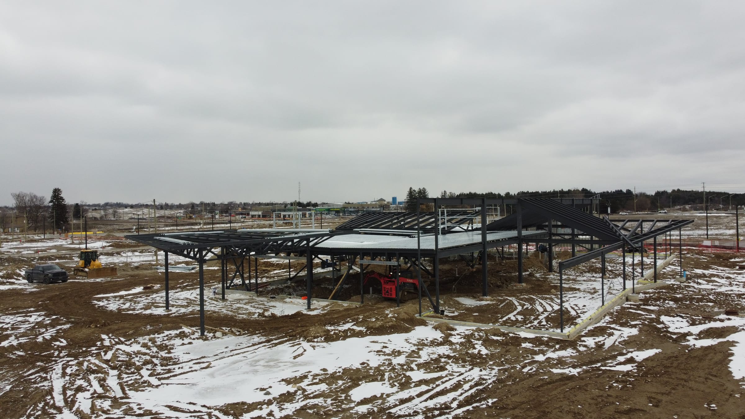 A wide aerial view of the whole steel frame on a snowy site, with vehicles and building materials around it.