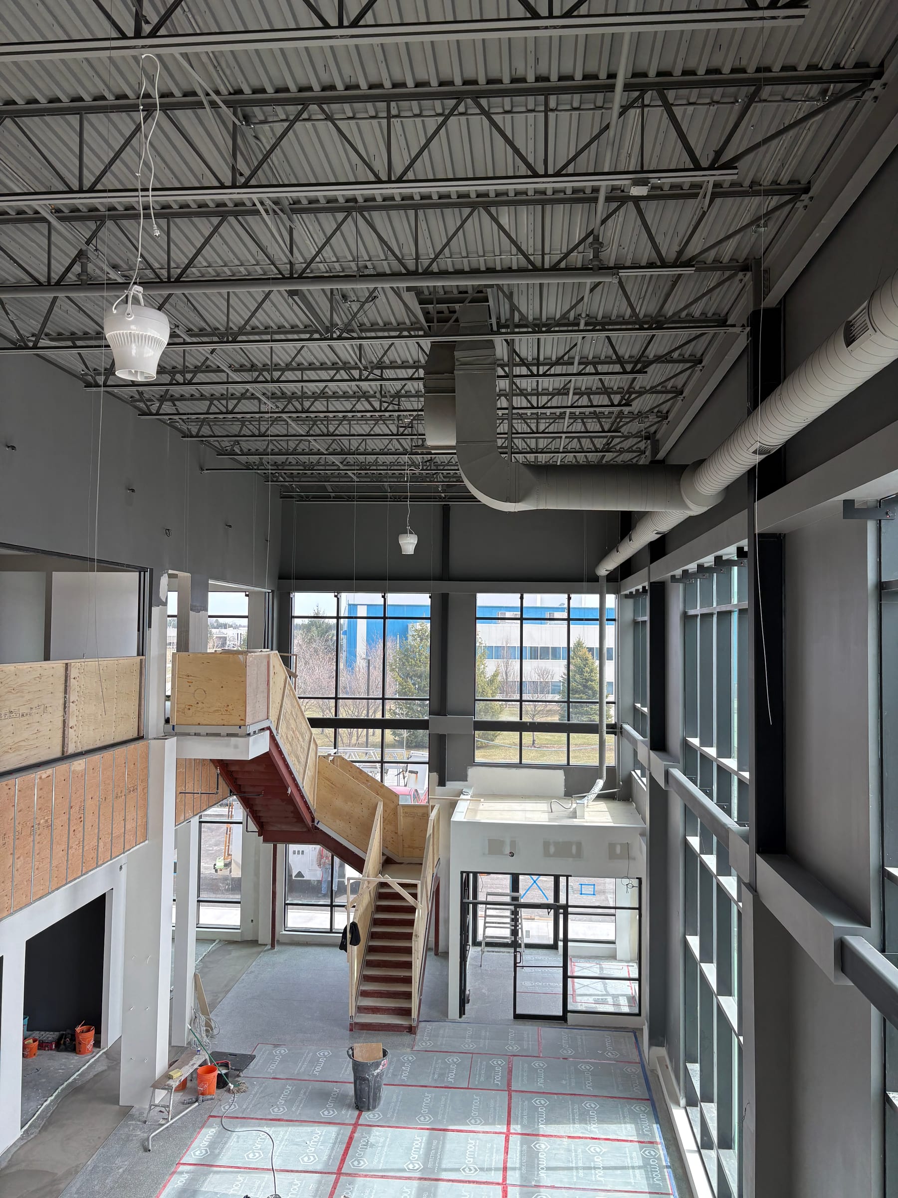 A two-storey office atrium under construction: a curtain wall, a steel stair boxed in plywood guards, floor protection, and exposed steel joists above.